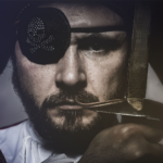 Man dressed as a pirate with a skull and cross-bones eye patch, holding a sword up in front of him with a gloved hand.