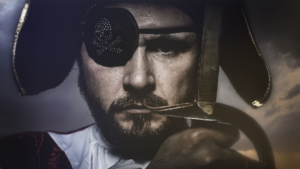 Man dressed as a pirate with a skull and cross-bones eye patch, holding a sword up in front of him with a gloved hand.