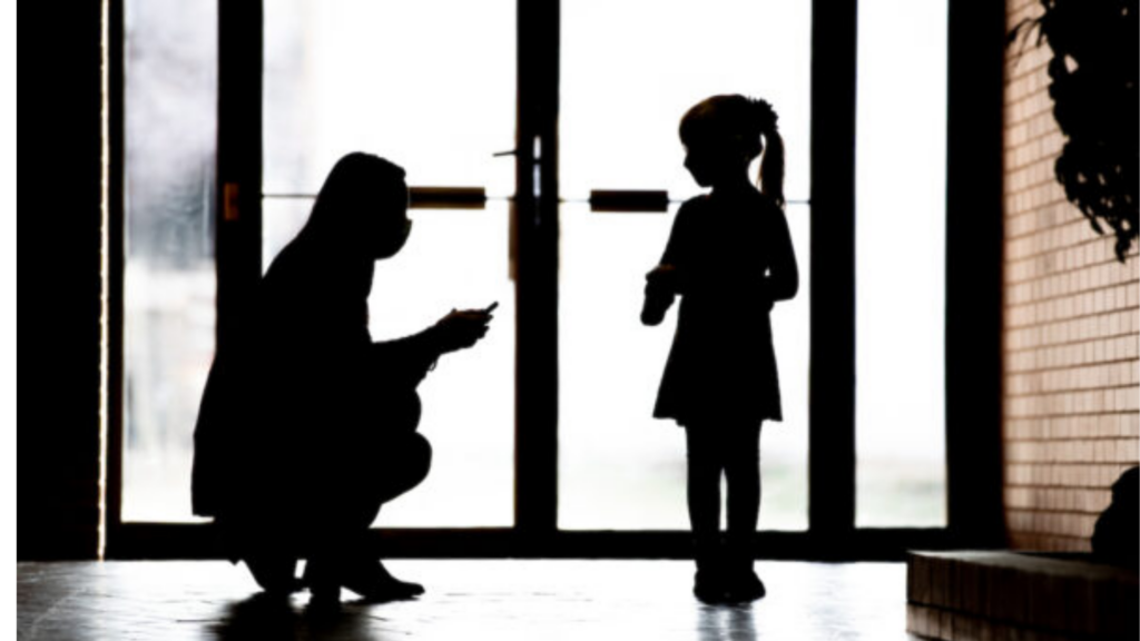 Woman kneels in shadow to speak with small girl by glass door