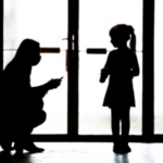 Woman kneels in shadow to speak with small girl by glass door