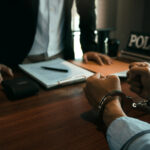 Police officer standing over desk intervewing handcuffed suspect