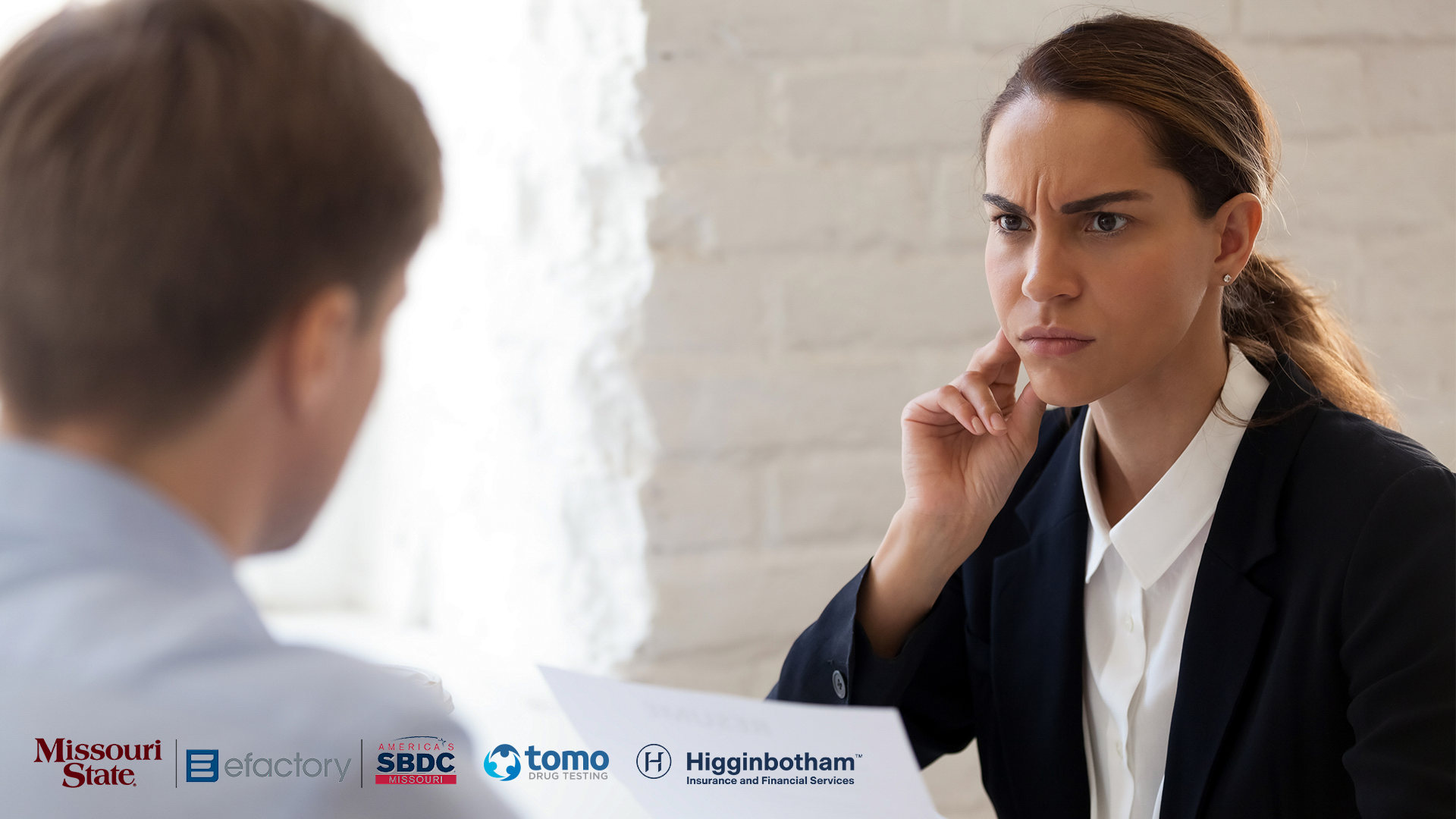 Business woman looking suspiciously at a man sitting across the table from her. The man's back is facing the camera and he is slightly blurred in the image.