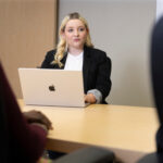 Blonde woman sitting across a conference table behind a laptop computer across the table from other people who are obscured from view.