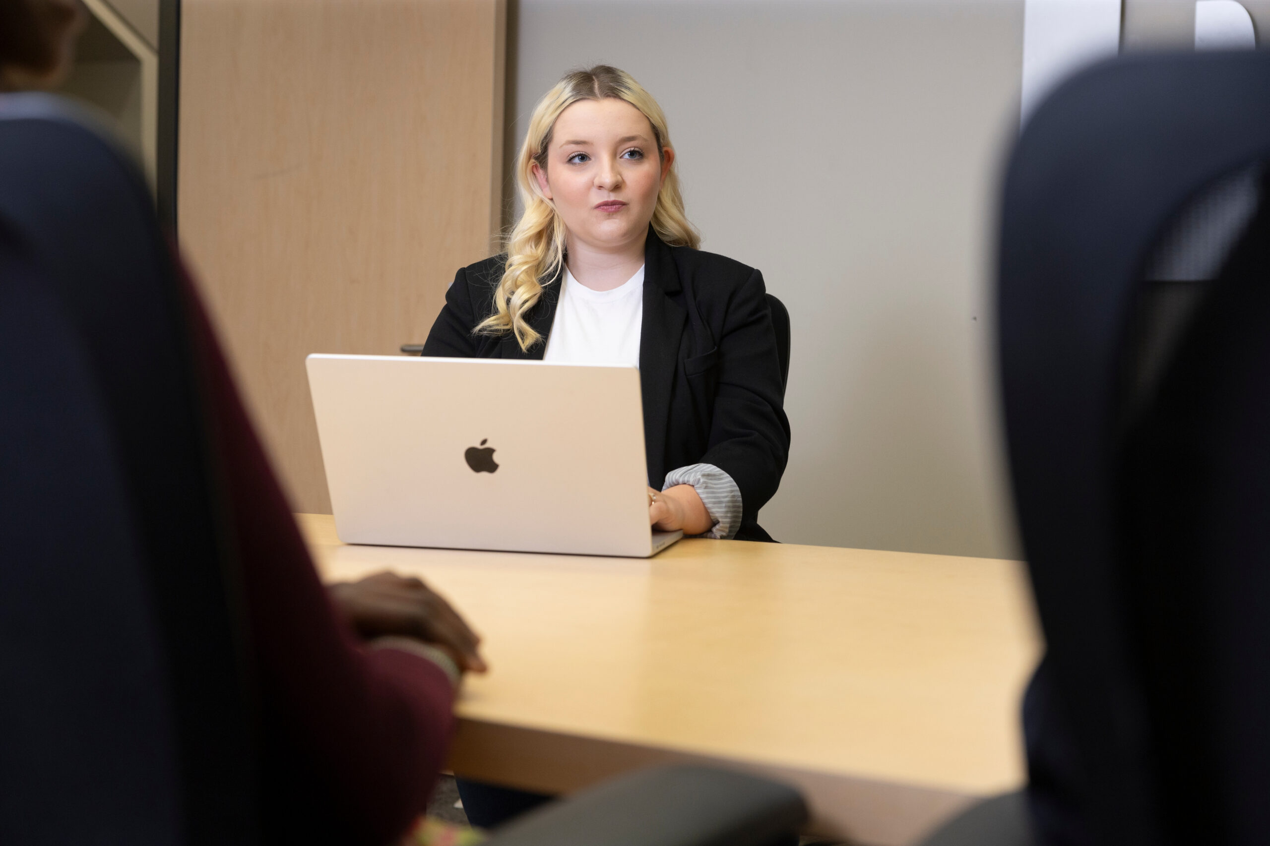 Blonde woman sitting across a conference table behind a laptop computer across the table from other people who are obscured from view.