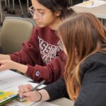 Two women seated at a table in front of a drawing of a business scorecard.