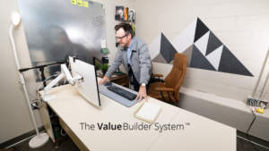 Businessman leaning over desk, looking at his computer screen with building blocks on wall resembling a mountain behind him.