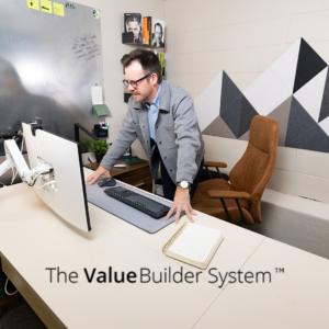 Businessman leaning over desk, looking at his computer screen with building blocks on wall resembling a mountain behind him.