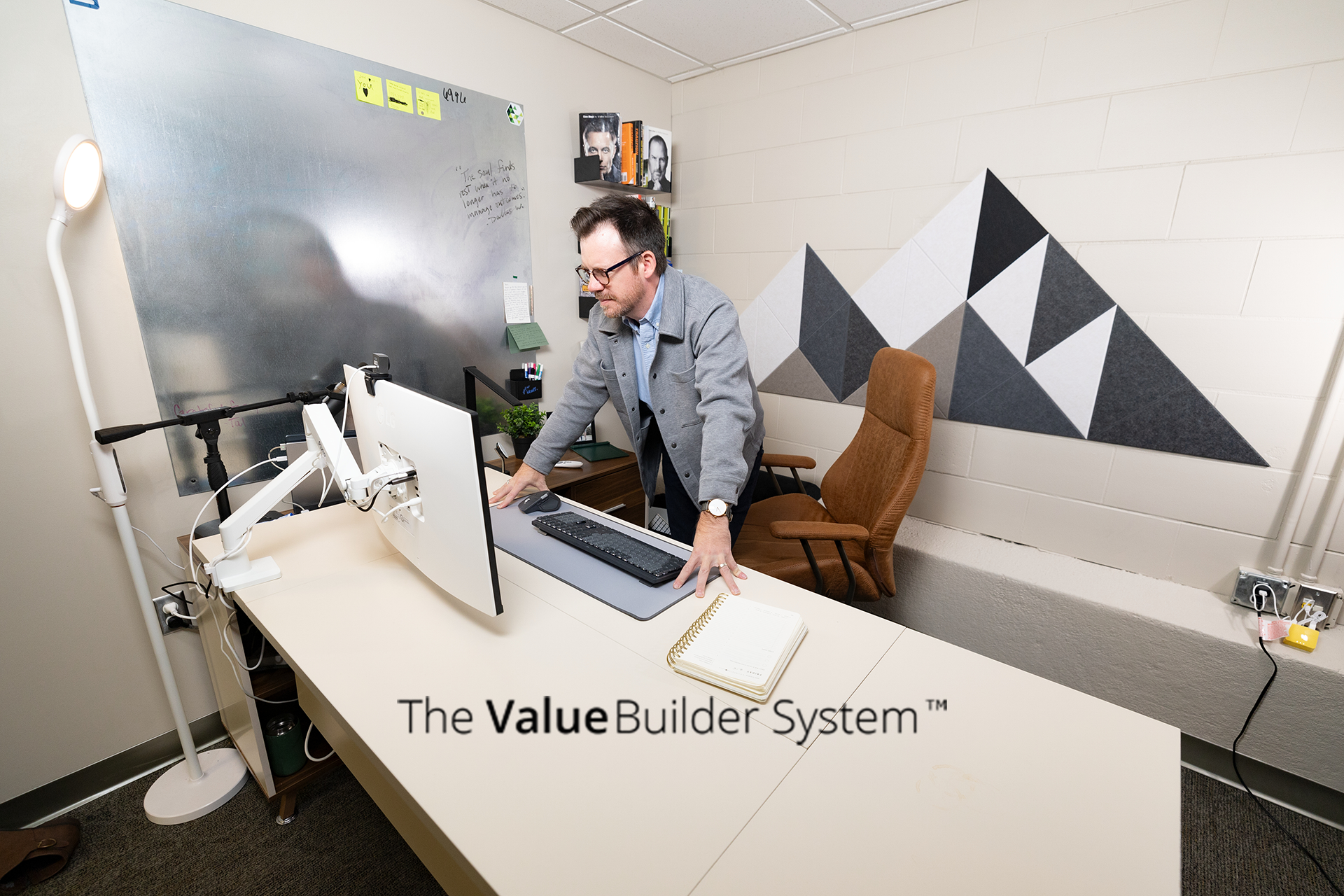 Businessman leaning over desk, looking at his computer screen with building blocks on wall resembling a mountain behind him.