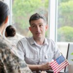 Female military member looking at a man seated in front of a laptop computer at a desk with two American flags displayed in a coffee cup.