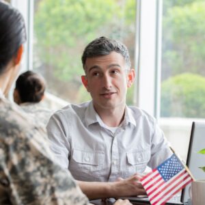 Female military member looking at a man seated in front of a laptop computer at a desk with two American flags displayed in a coffee cup.
