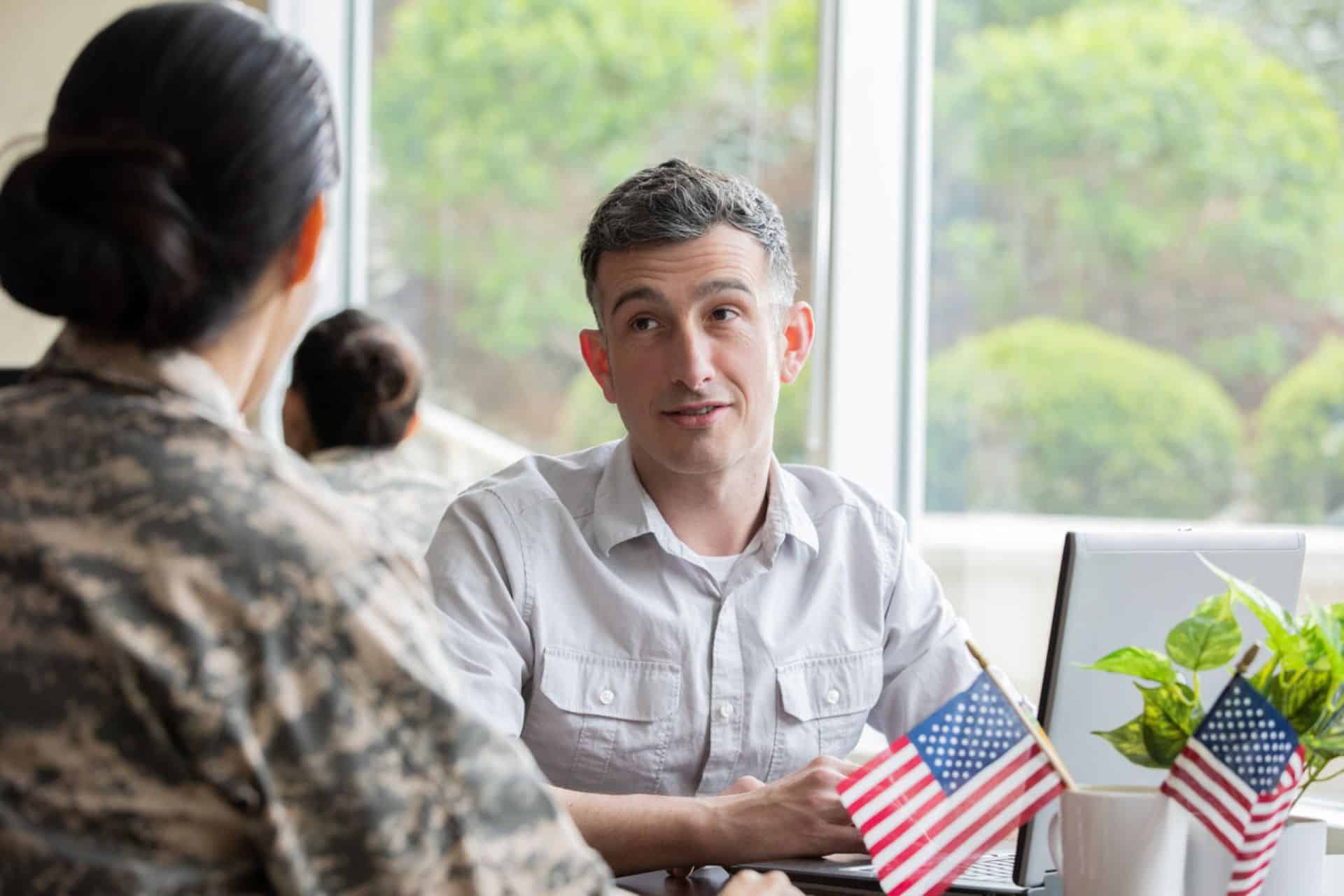 Female military member looking at a man seated in front of a laptop computer at a desk with two American flags displayed in a coffee cup.